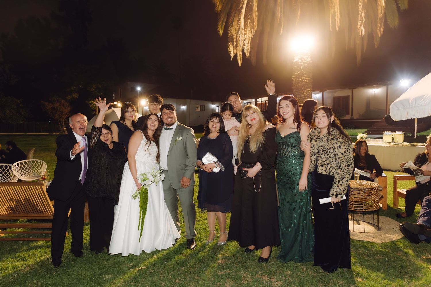 Large group of wedding guests posing with bride and groom at outdoor evening reception.
