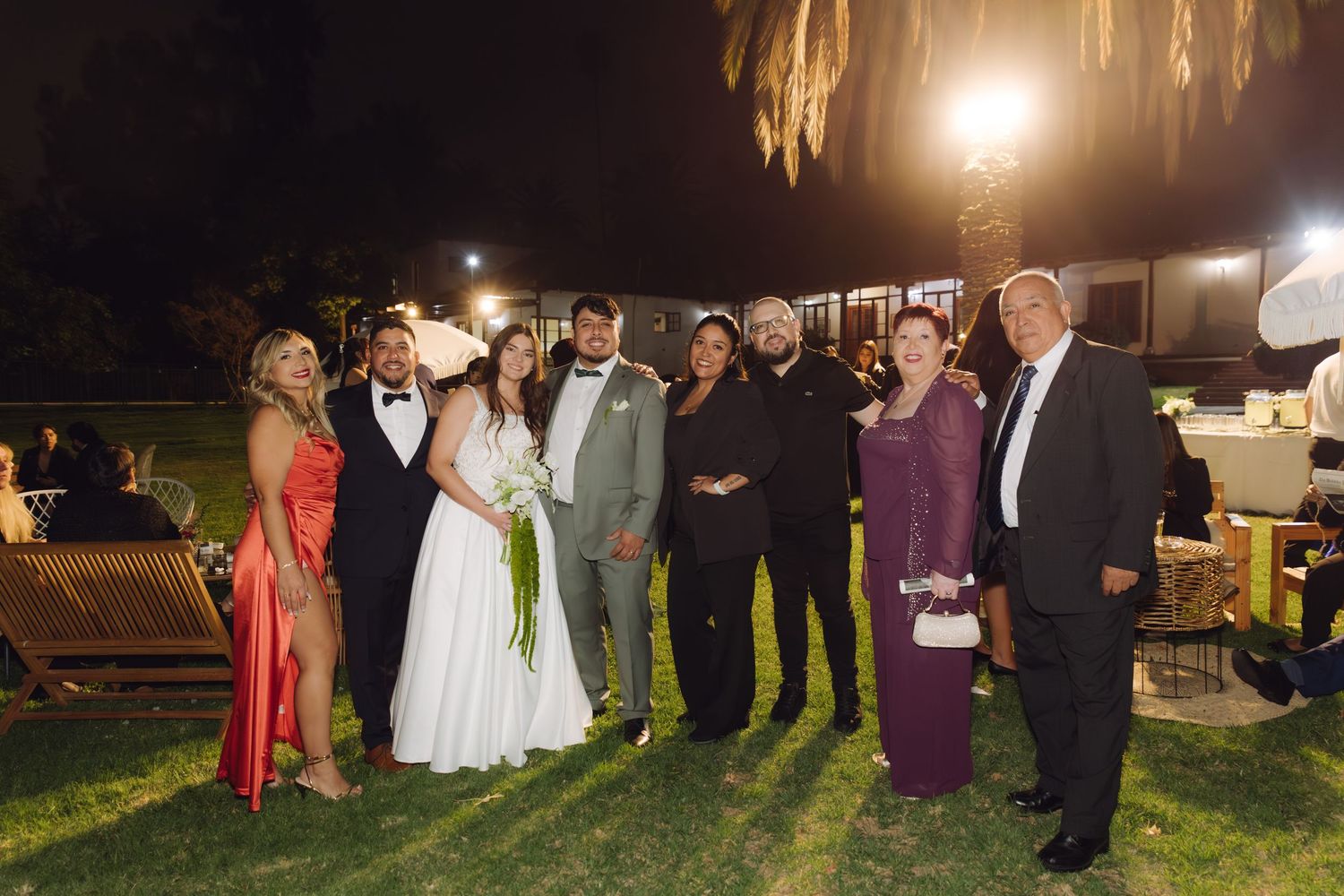 Bride and groom surrounded by elegantly dressed family members posing at nighttime wedding reception.