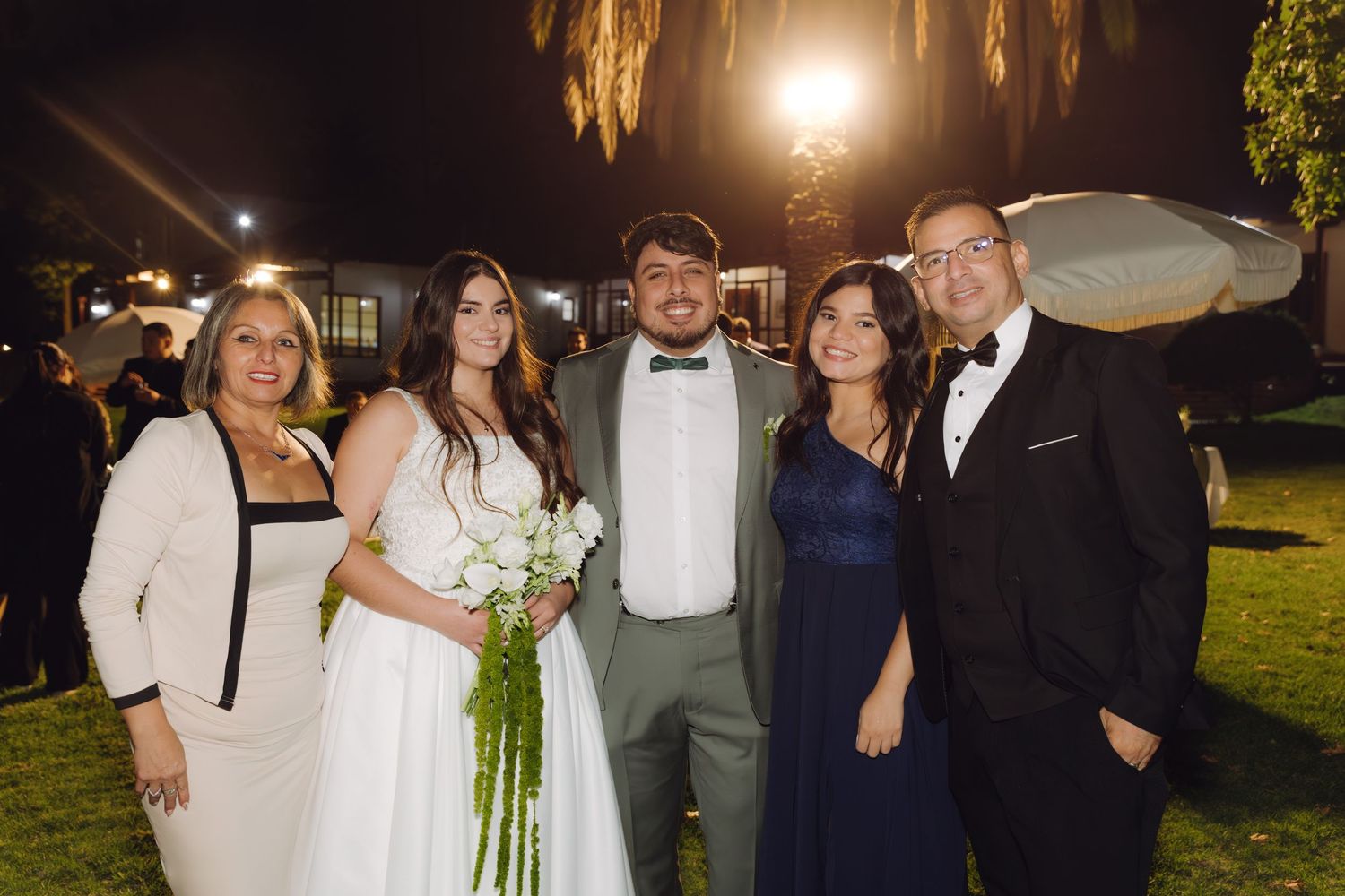 Bride and groom pose with four family members smiling at outdoor evening wedding celebration.