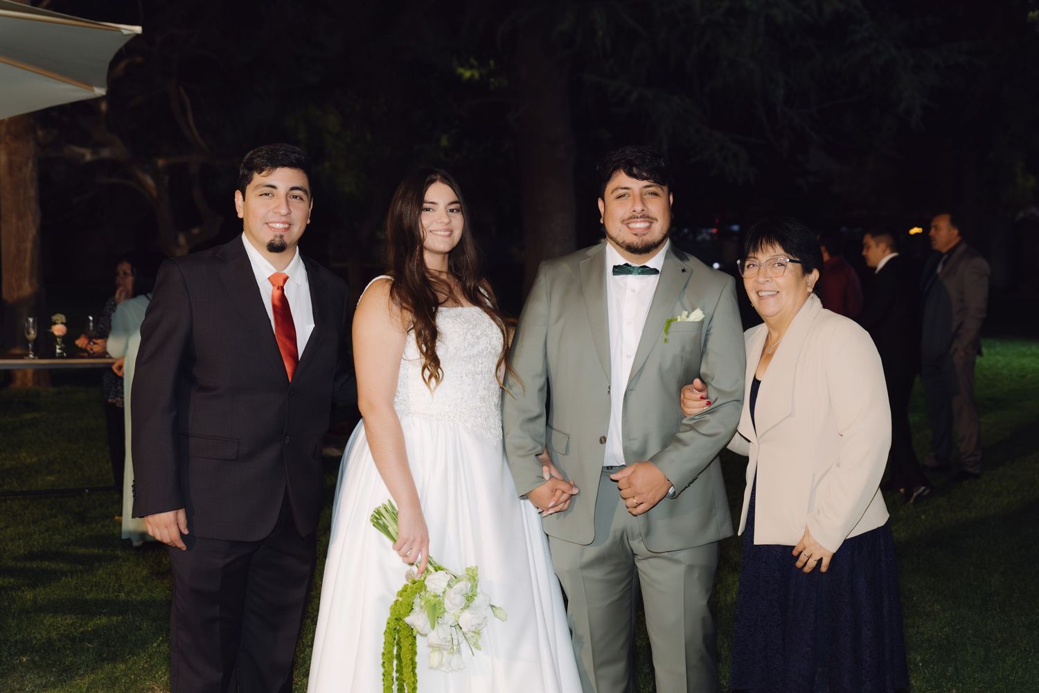 Bride and groom pose with two family members under trees at outdoor evening wedding.