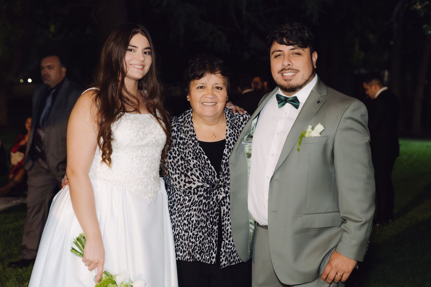 Bride and groom smile with older woman guest at outdoor nighttime wedding reception.