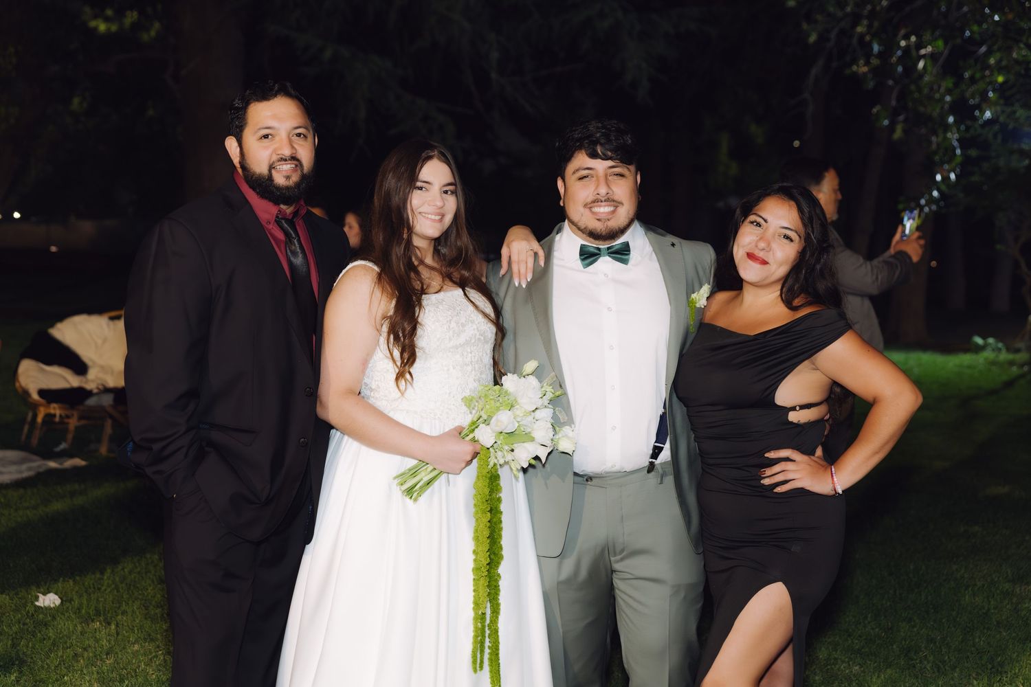 Wedding party of four posing outdoors at night, featuring bride in white gown with green bouquet and groom in gray suit.