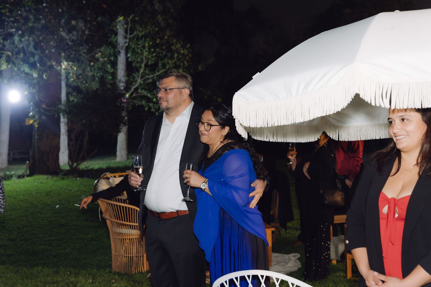 Three guests standing outdoors at night near a white umbrella at a wedding reception event.