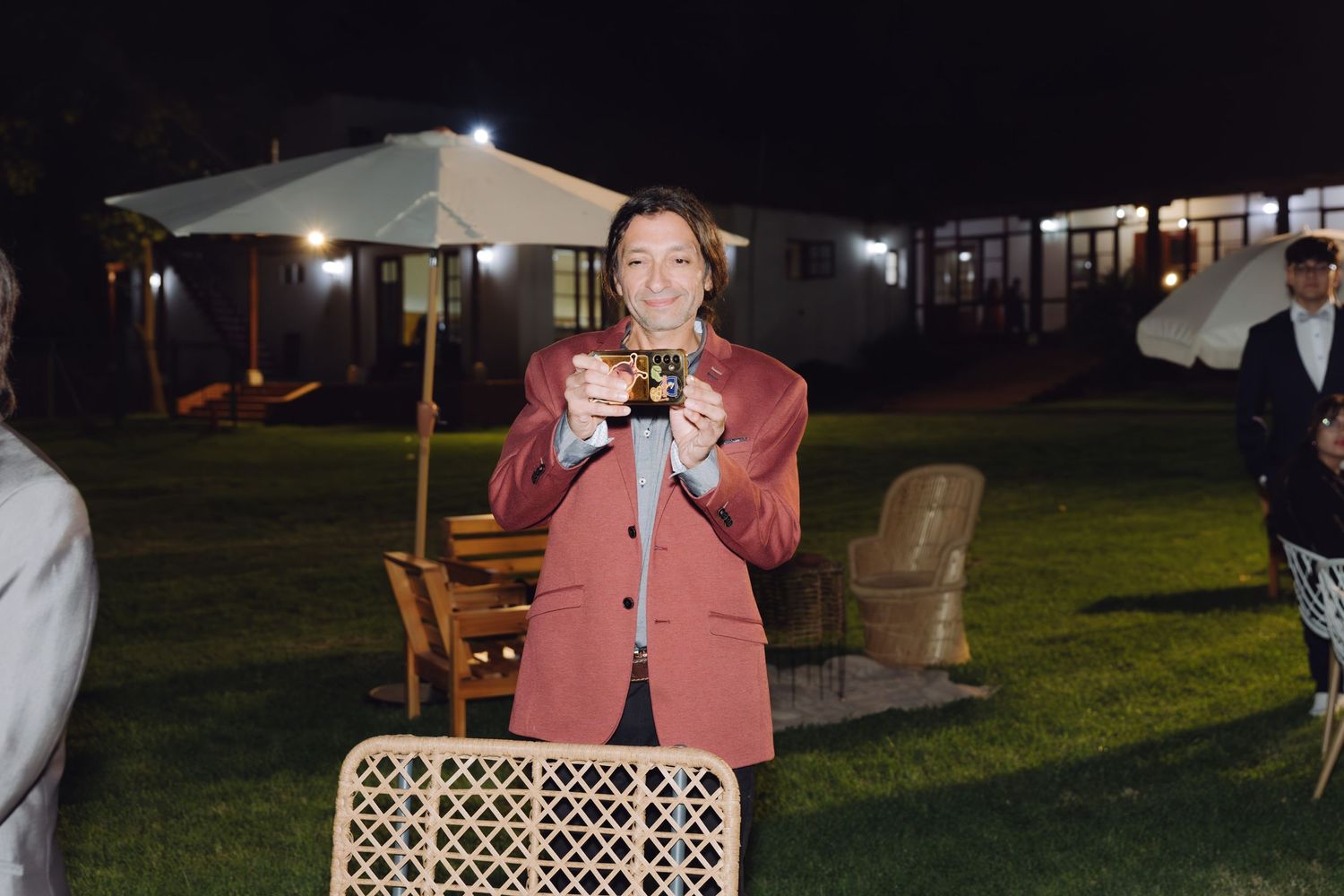 Woman in rose blazer smiling and holding a cup at an outdoor nighttime wedding reception.