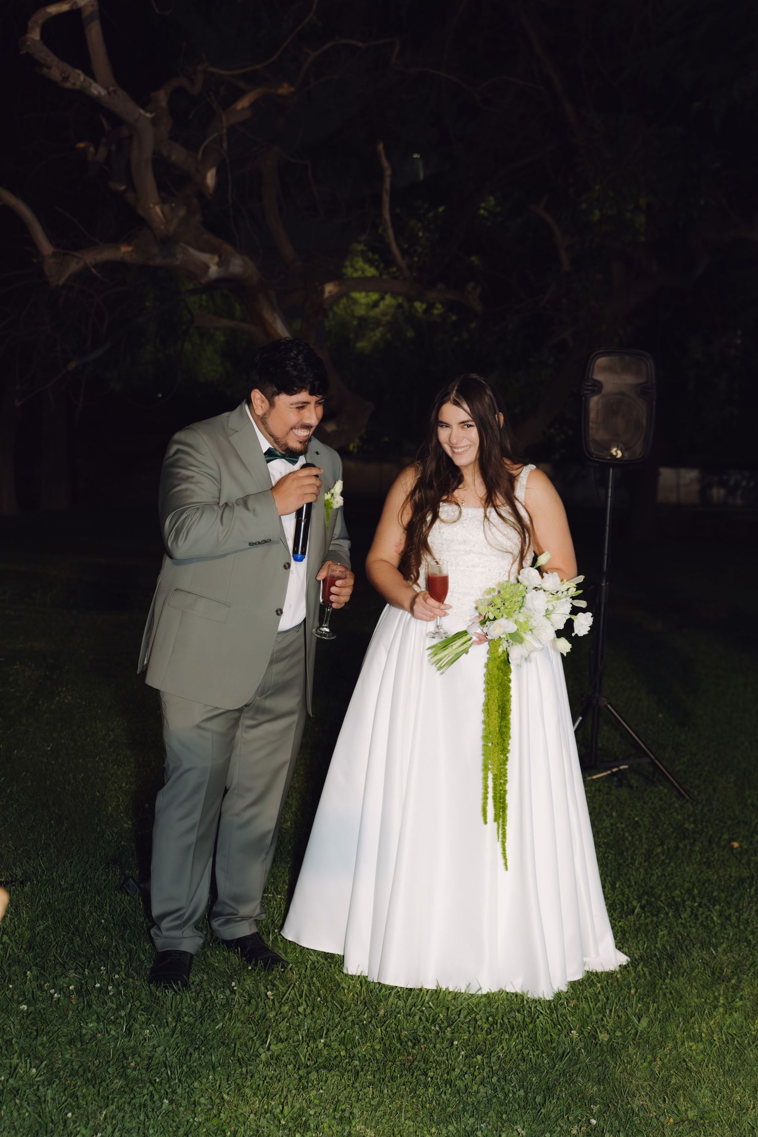 Bride in white gown holding bouquet stands beside groom in gray suit speaking into microphone outdoors.