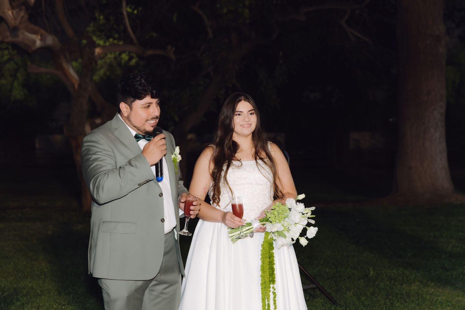 Bride holding white bouquet and groom speaking into microphone at outdoor nighttime wedding ceremony.