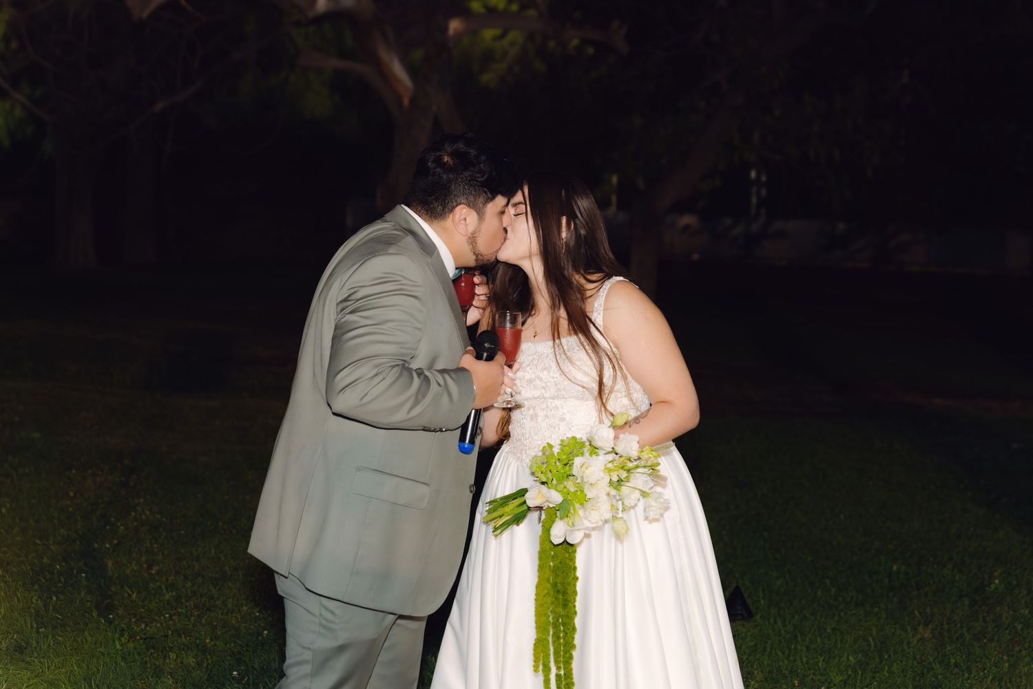 Bride and groom sharing a kiss outdoors at night, bride holding white and green floral bouquet.