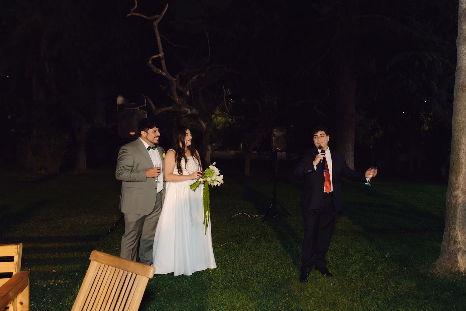 Bride and groom standing outdoors at night as a guest in red tie approaches them at wedding.