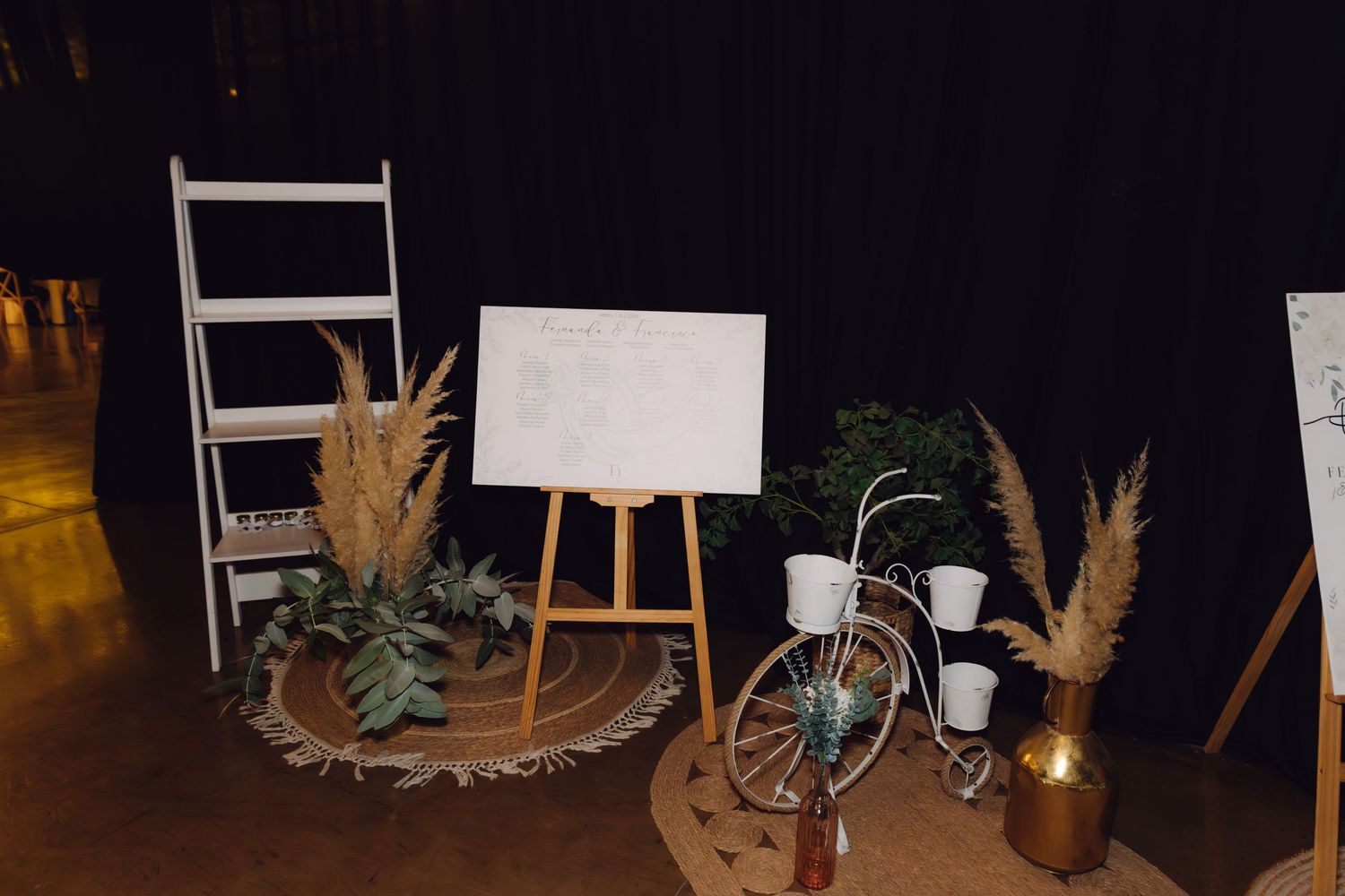 Rustic boho wedding welcome display with pampas grass, white easel sign, decorative bicycle, and ladder shelf on jute rug.