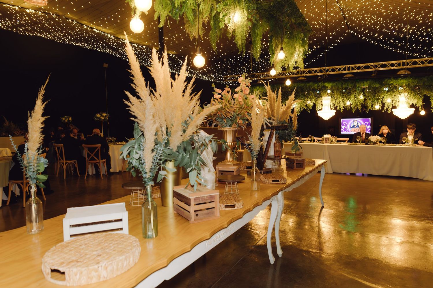 Elegant outdoor wedding reception with long decorated table, pampas grass centerpieces, string lights, and lush green canopy.