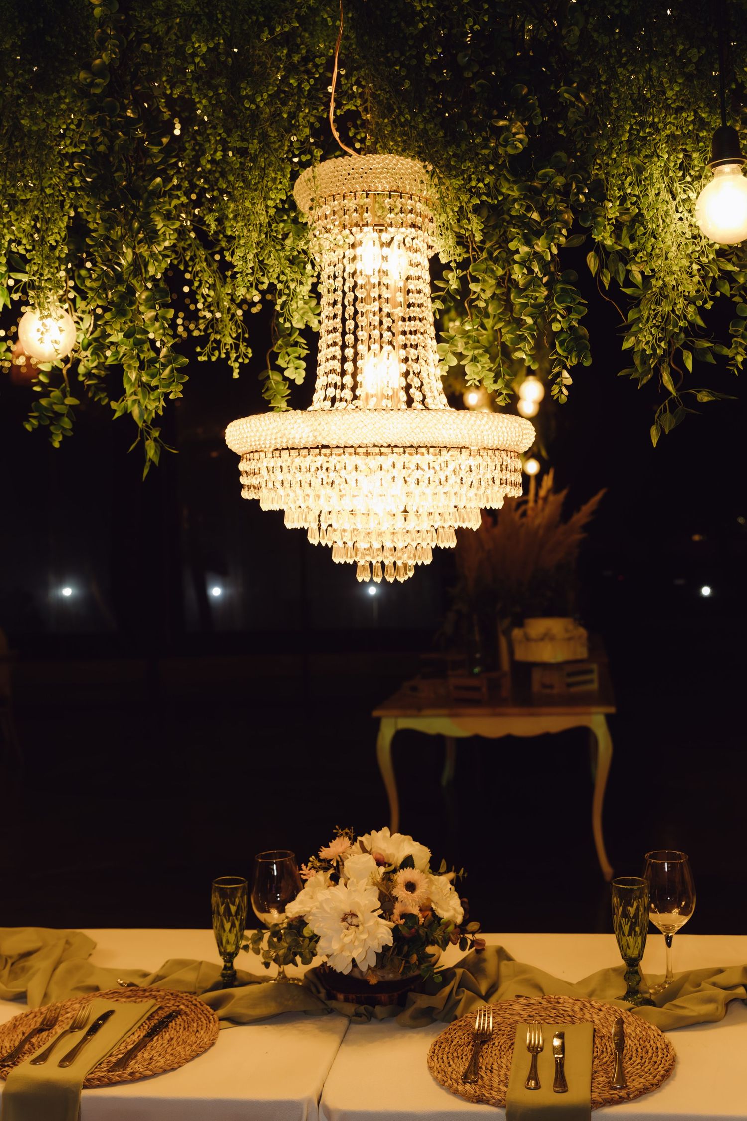 Stunning crystal chandelier illuminating a romantic wedding table setting with white flowers and greenery overhead at night.