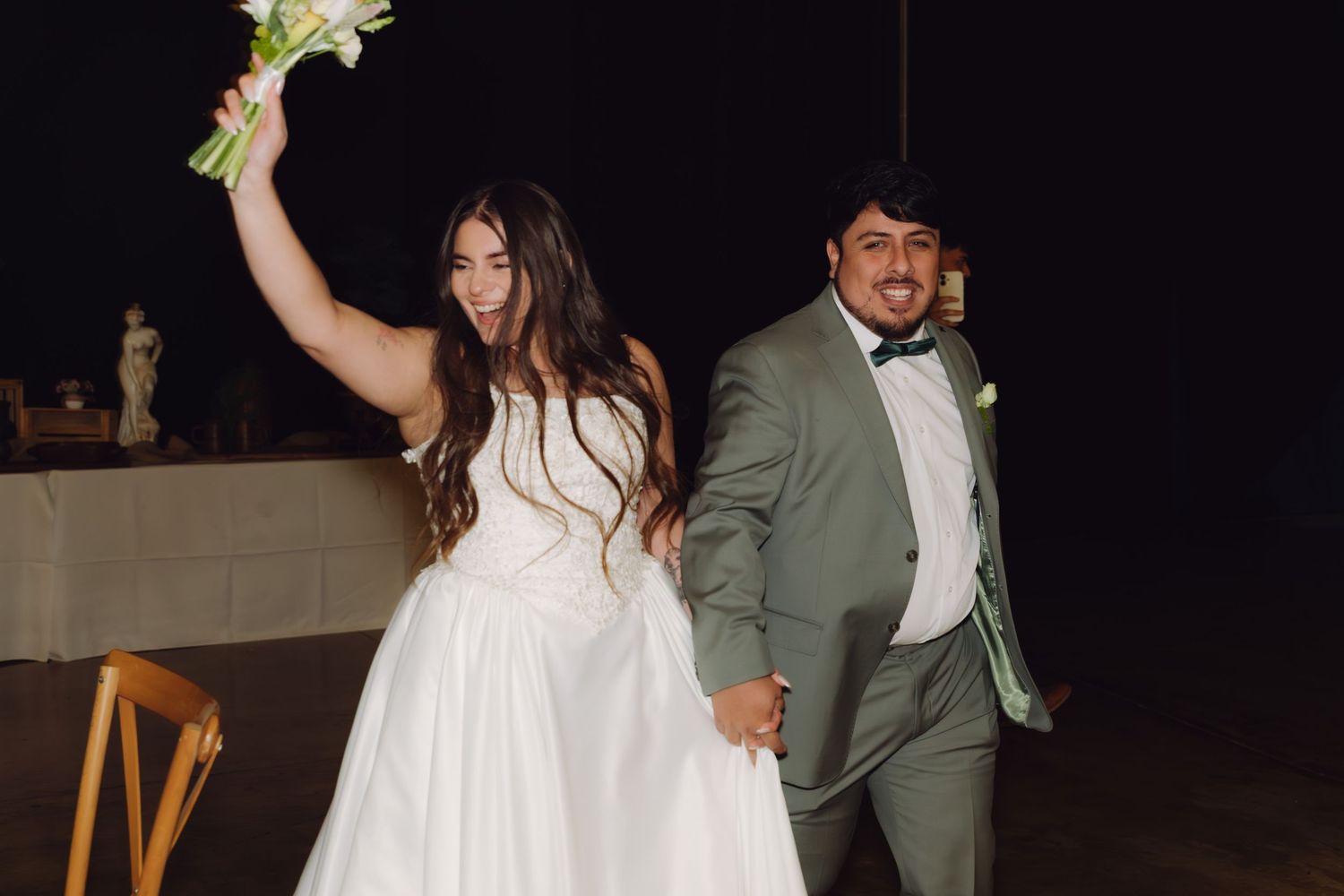 Joyful newlyweds making grand entrance, bride in white gown raising bouquet, groom in sage green tuxedo, celebrating together.
