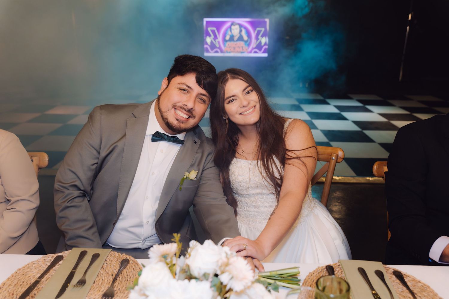 Smiling bride and groom seated at wedding reception sweetheart table with white floral centerpiece and woven placemats.