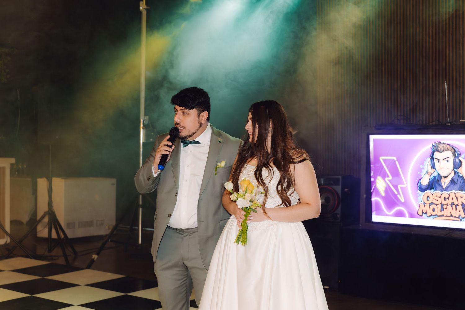 Groom speaking into microphone on checkered dance floor beside bride holding bouquet with colorful stage lighting behind them.