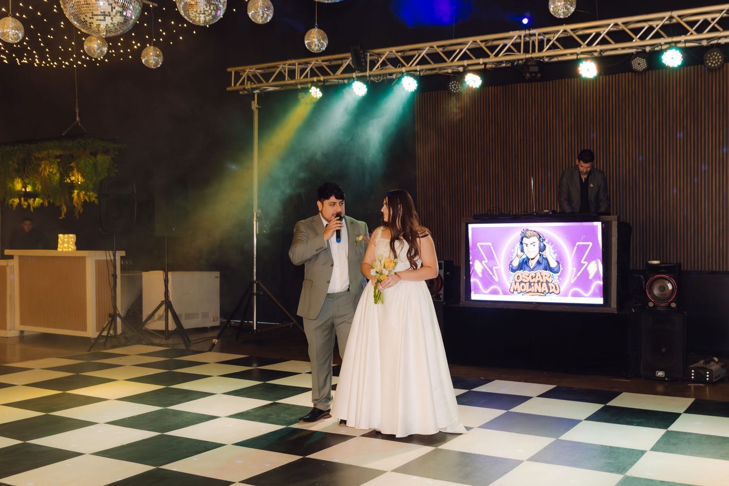 Bride and groom on black and white checkered dance floor with DJ booth, disco balls, and vibrant stage lighting.