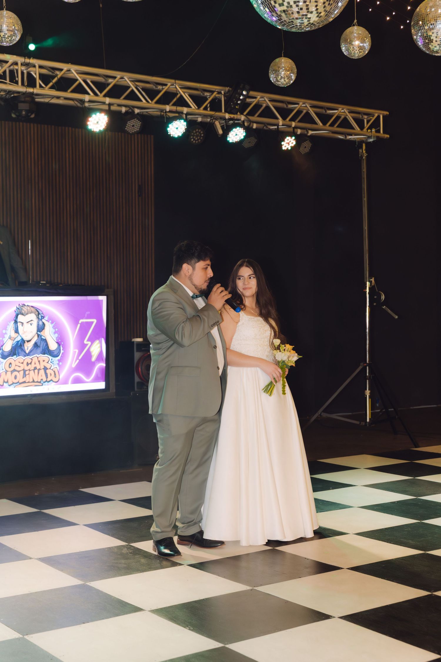 Elegant couple standing on checkered ballroom floor, groom speaking into microphone beside bride holding white bouquet at reception.
