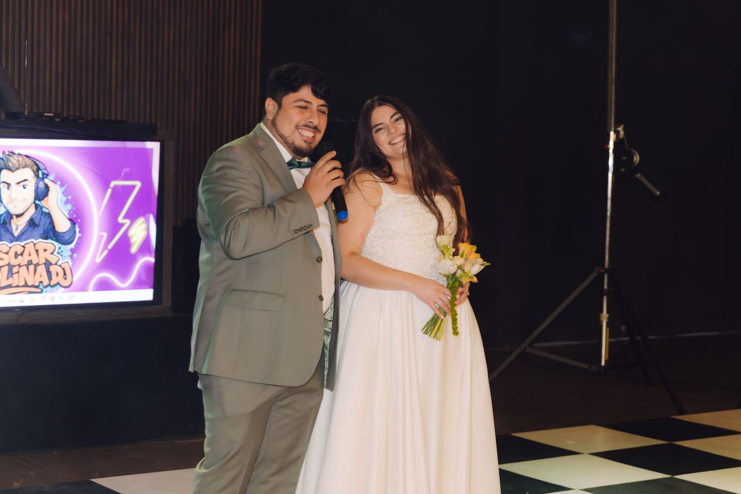Groom addressing wedding guests with microphone while bride stands beside him holding colorful bouquet on dance floor.