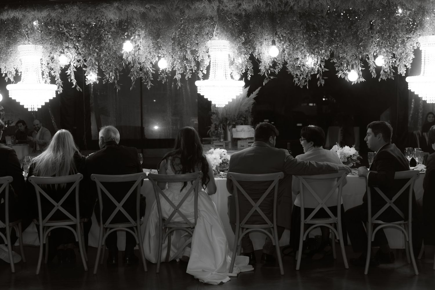 Black and white photo of wedding party seated at long head table under glowing chandeliers and floral canopy.