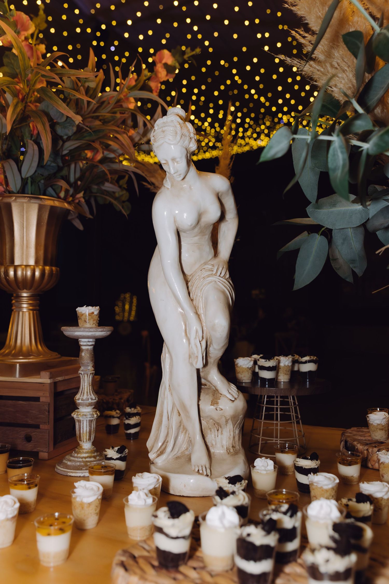 Wedding dessert table with white goddess statue centerpiece surrounded by assorted mini dessert cups and fairy lights.