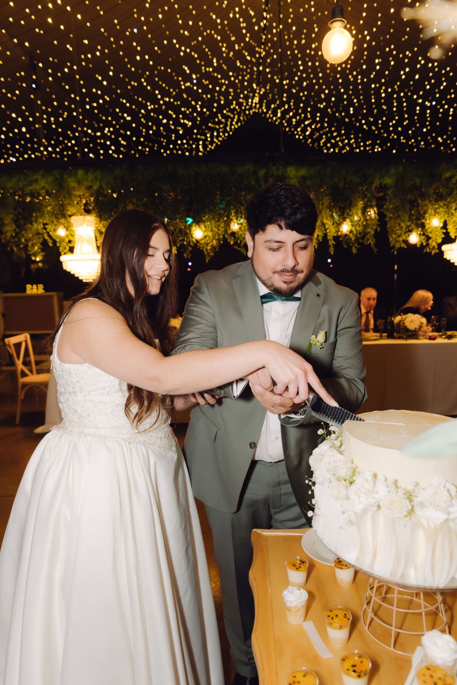 Bride and groom cutting their white floral wedding cake under a canopy of fairy lights at reception.