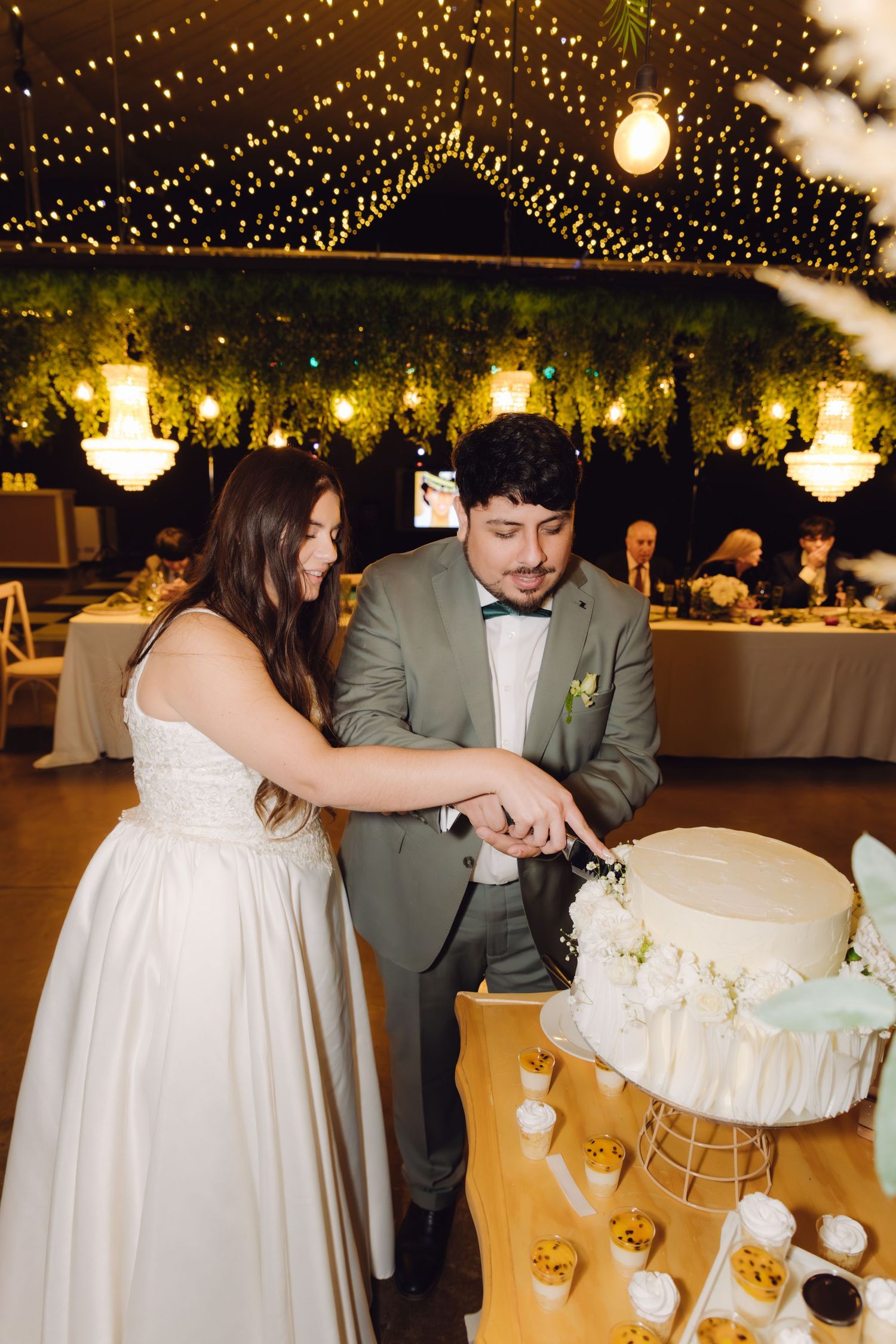 Newlyweds cutting their elegant white wedding cake together under warm fairy lights at their reception venue.