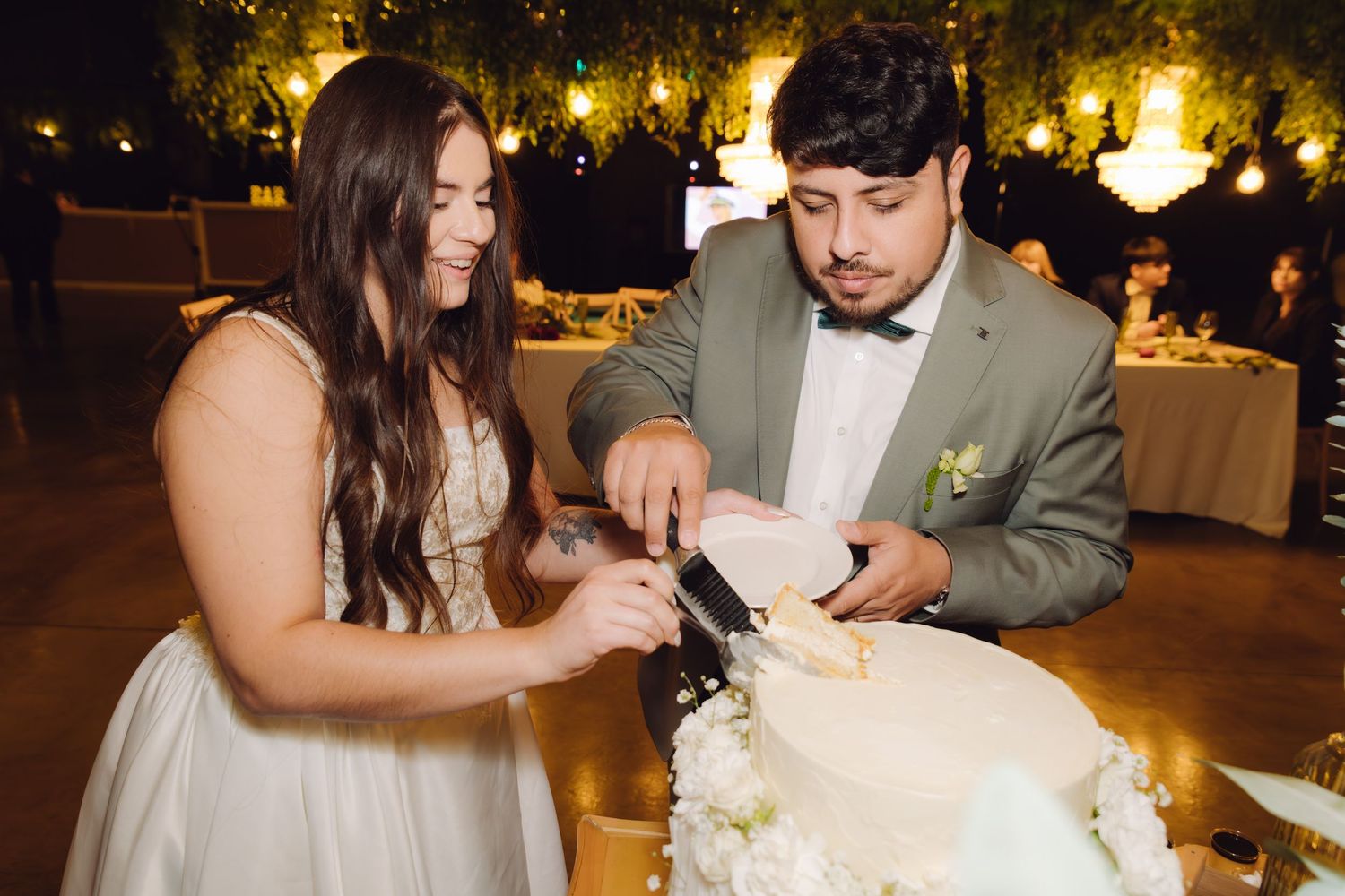 Bride and groom cutting their white wedding cake together at an elegant reception with warm string lights.