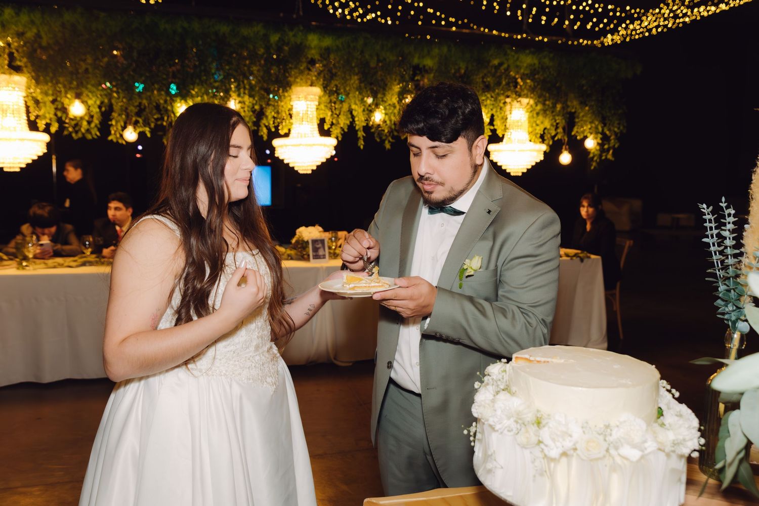 Newlyweds sharing a piece of wedding cake at their romantic reception venue decorated with fairy lights.