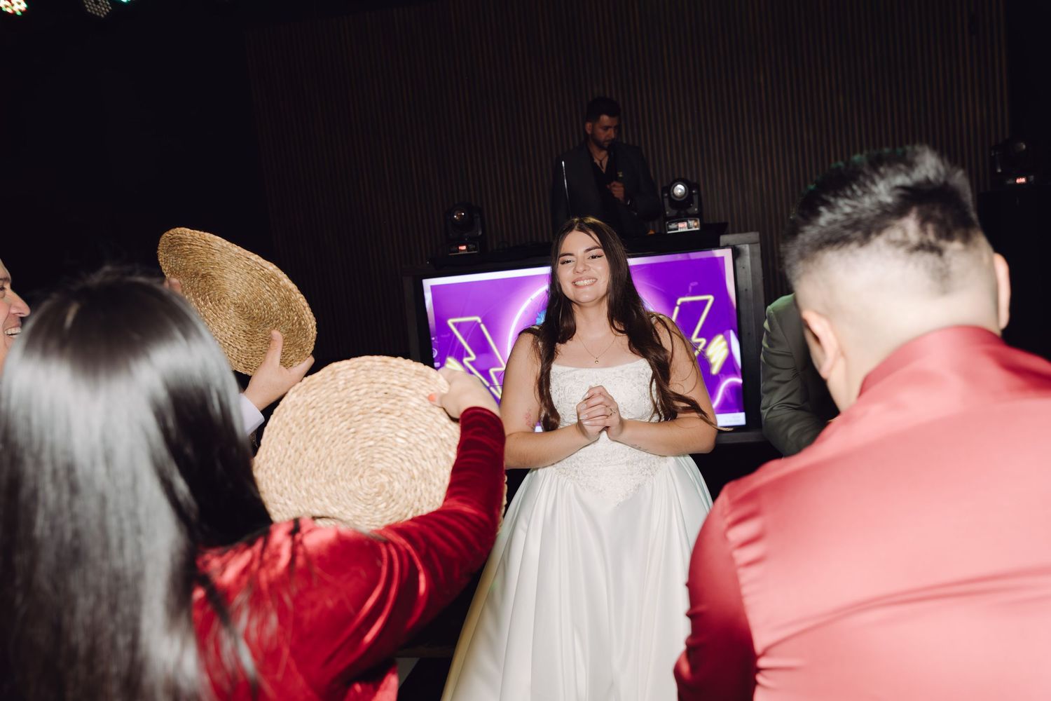 Bride in white gown surrounded by guests with hats on a dark wedding reception dance floor.
