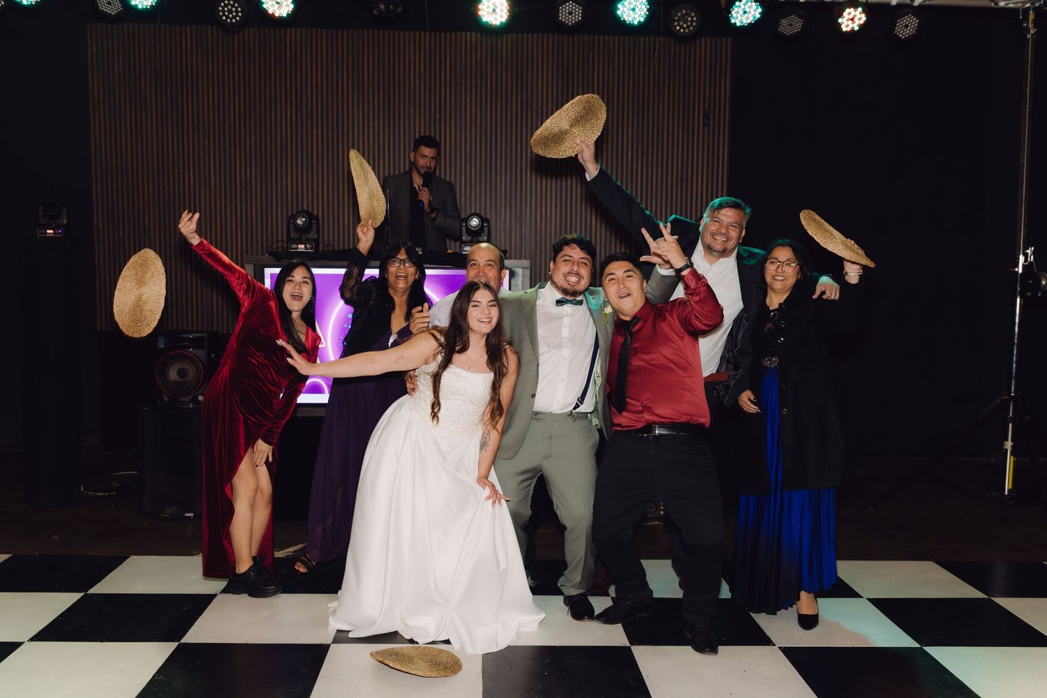 Wedding party group photo on checkered dance floor with hats raised in celebration at reception.
