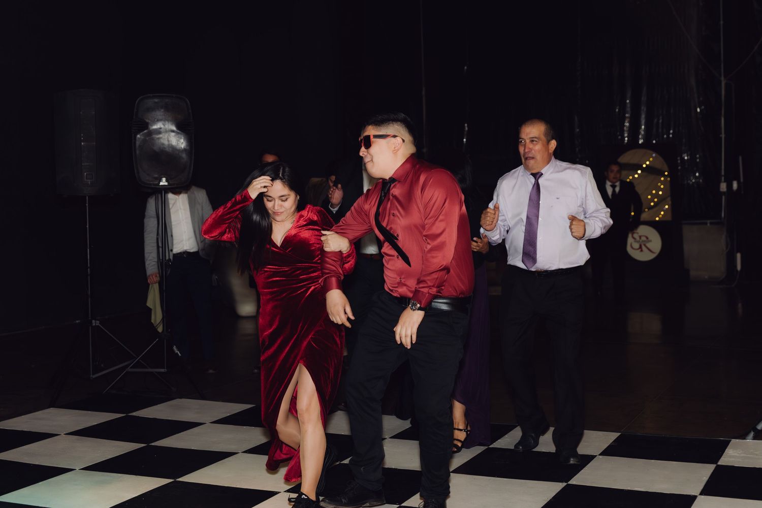 Guests in red velvet and burgundy outfits dancing on checkered floor at wedding reception.