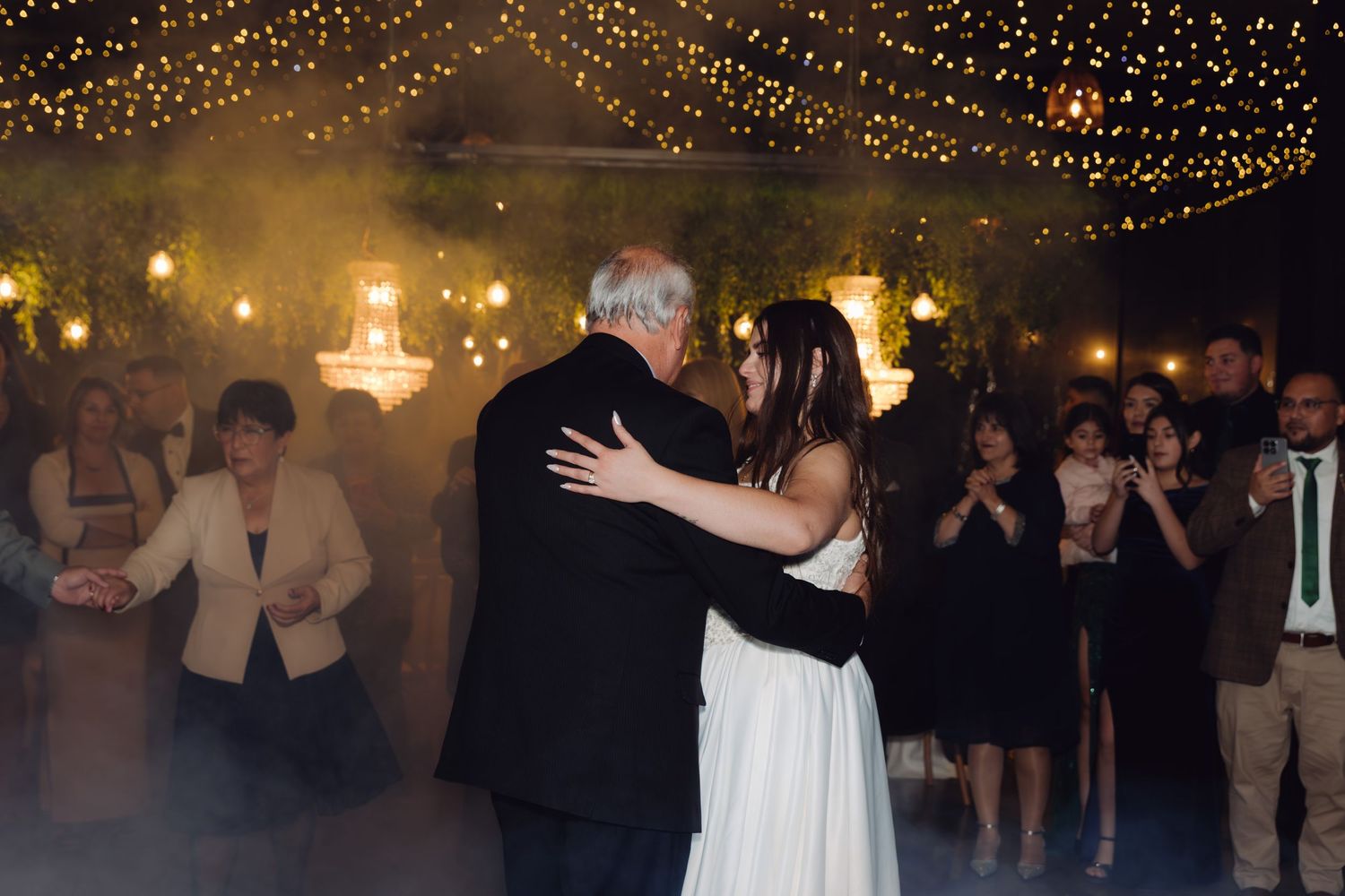 Bride and father share emotional first dance under warm fairy lights with fog effect at wedding reception.