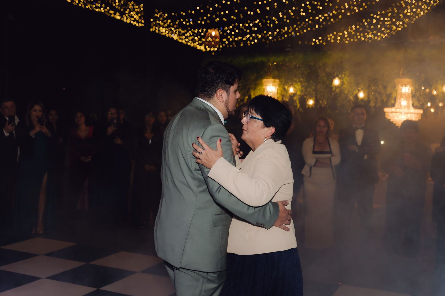 Groom shares tender mother-son dance under twinkling fairy lights at elegant wedding reception.