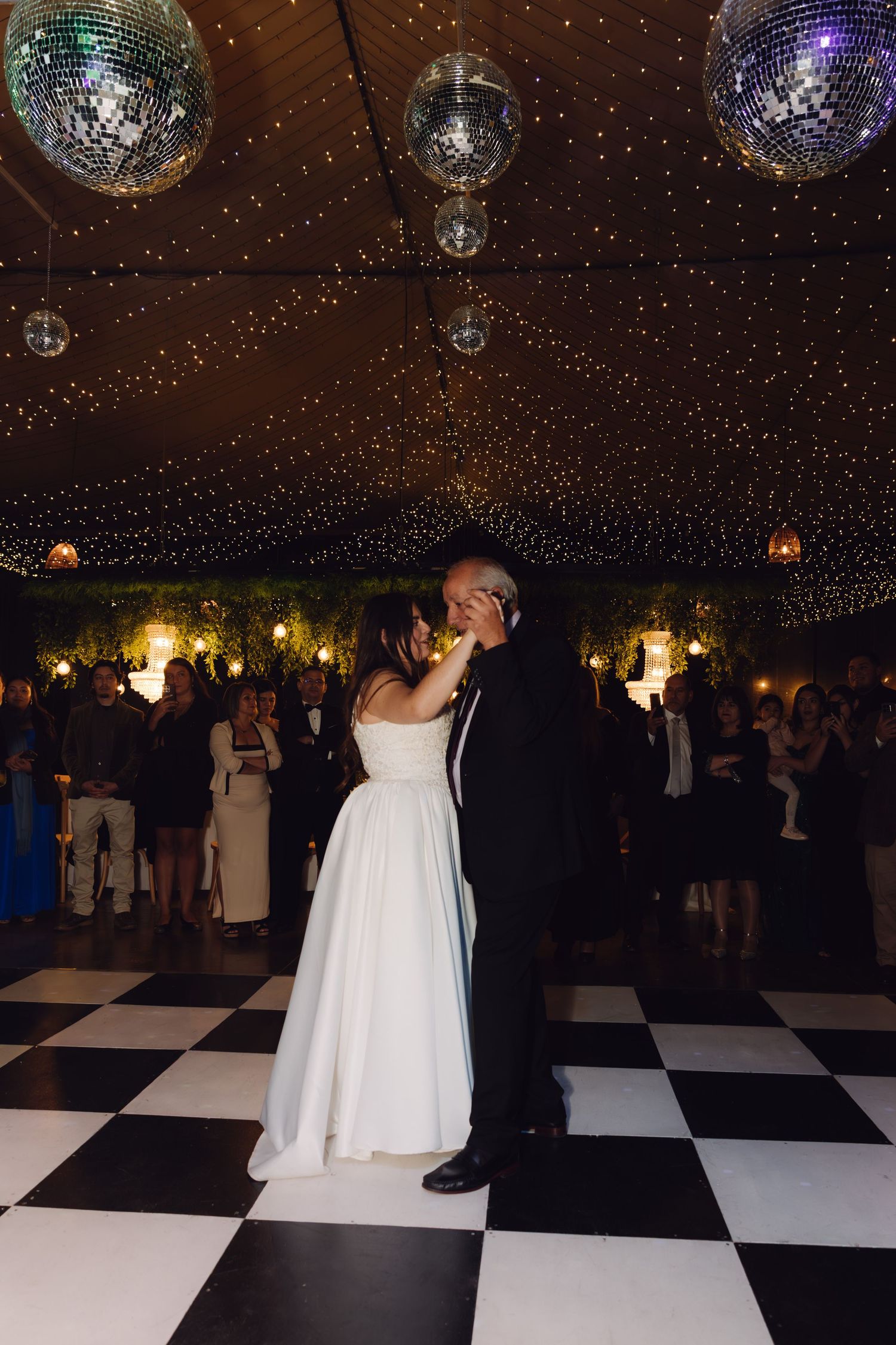 Bride and father share emotional dance under disco balls and twinkling lights on checkered dance floor.