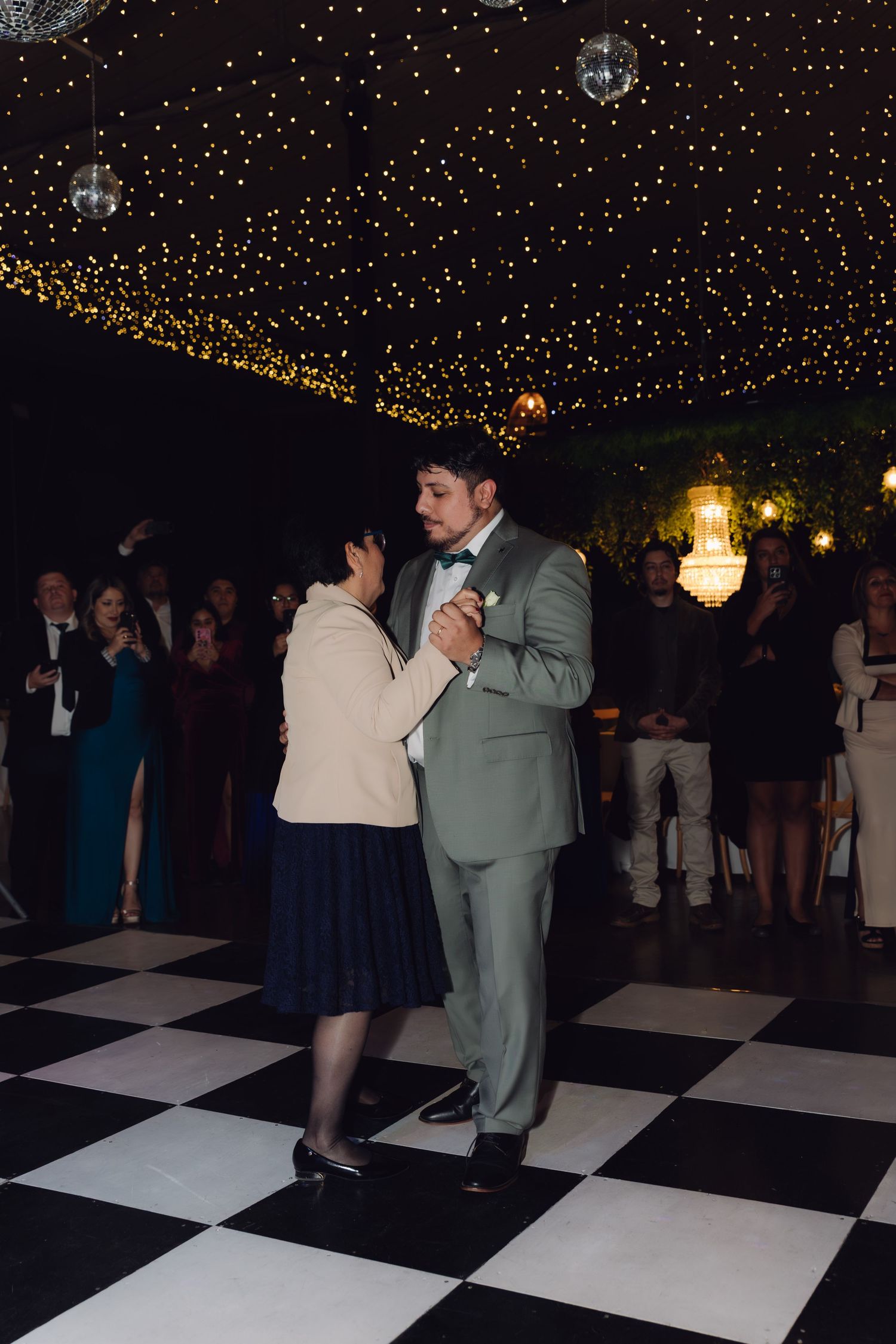Groom in sage suit dances with mother on black and white checkered floor under starlit ceiling.