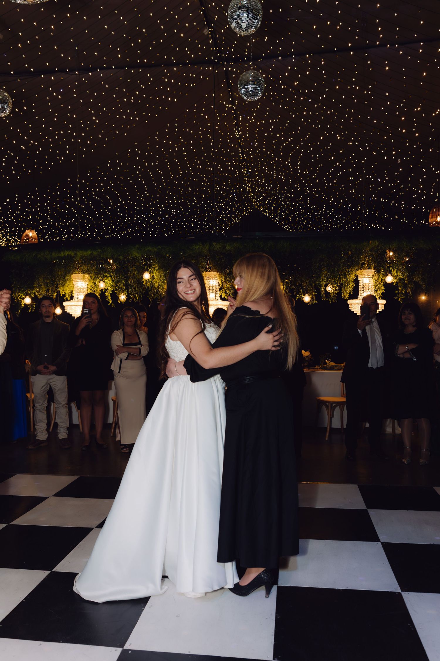 Smiling bride in white gown shares heartfelt dance with woman in black dress at wedding reception.