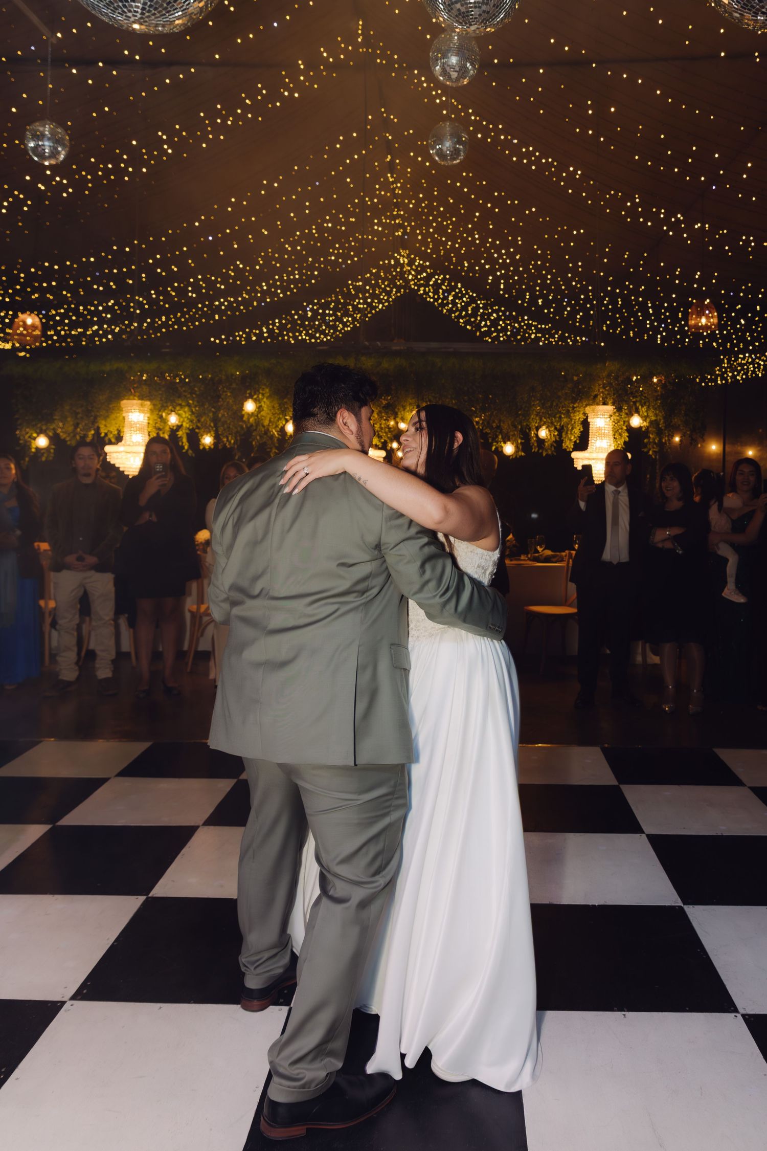 Bride and groom share romantic first dance under sparkling fairy lights on checkered ballroom floor.
