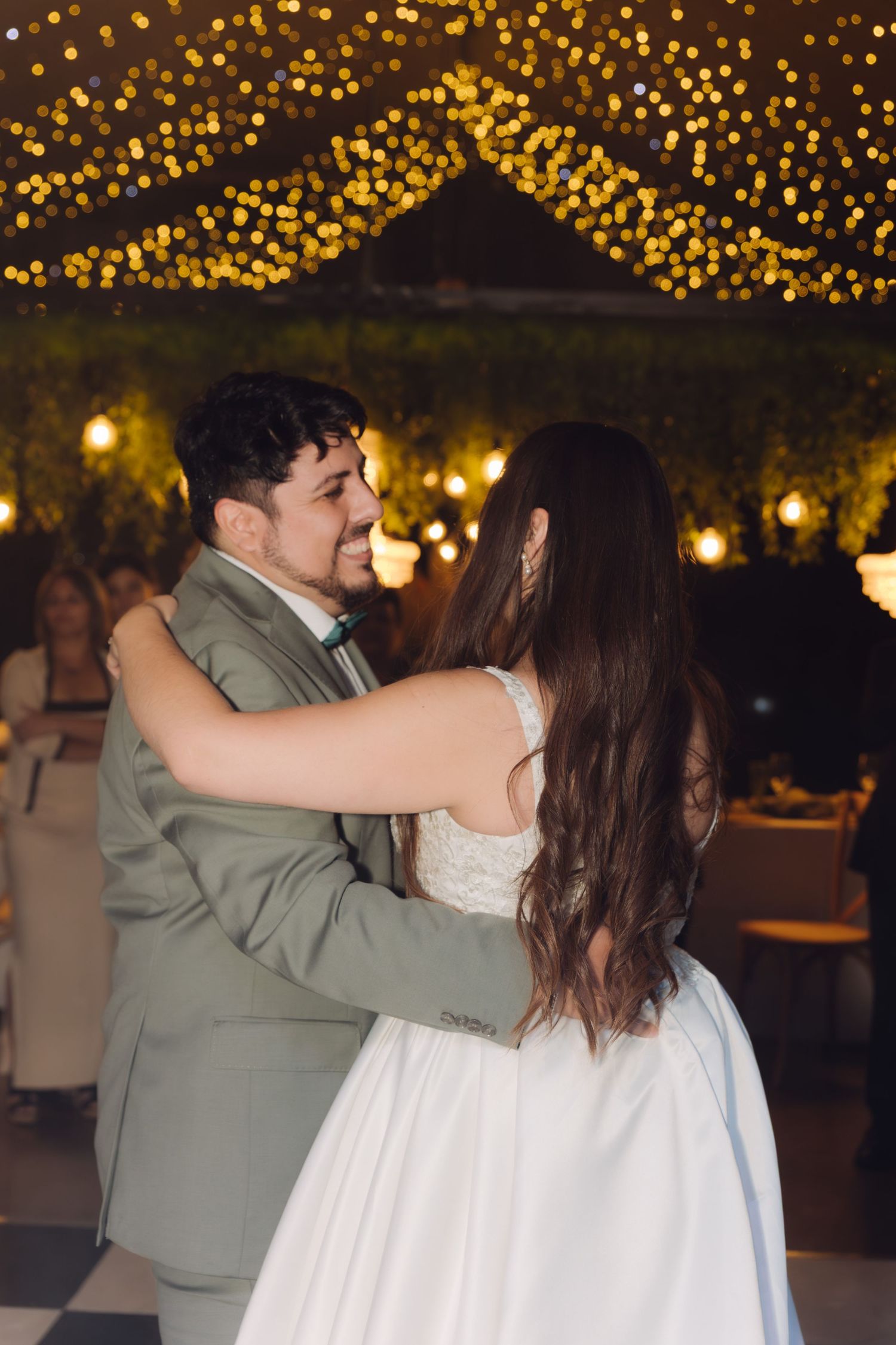 Smiling groom in sage suit gazes at bride during first dance under glowing string lights.