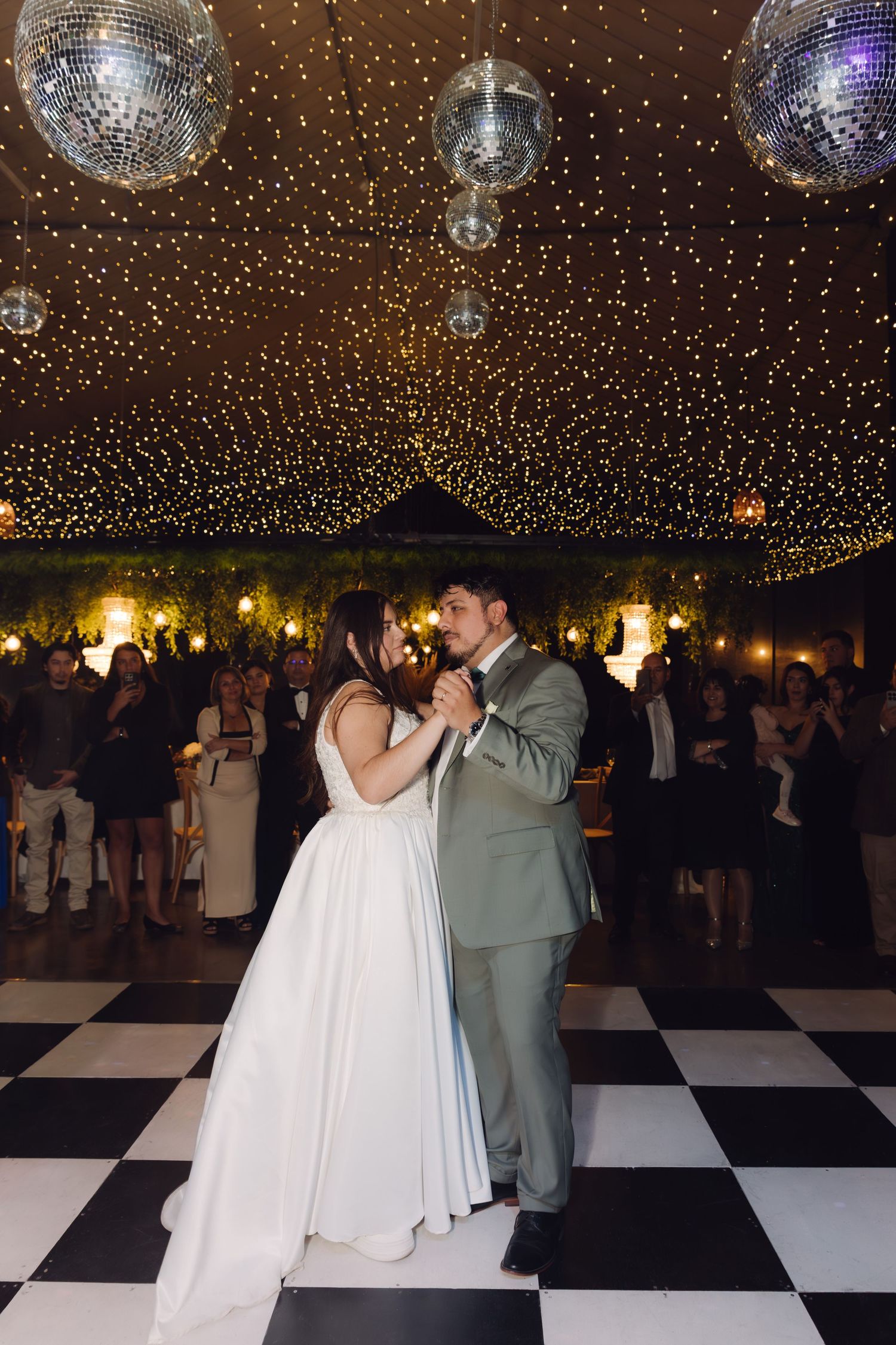 Newlyweds share first dance under disco balls and starlit ceiling on classic checkered dance floor.