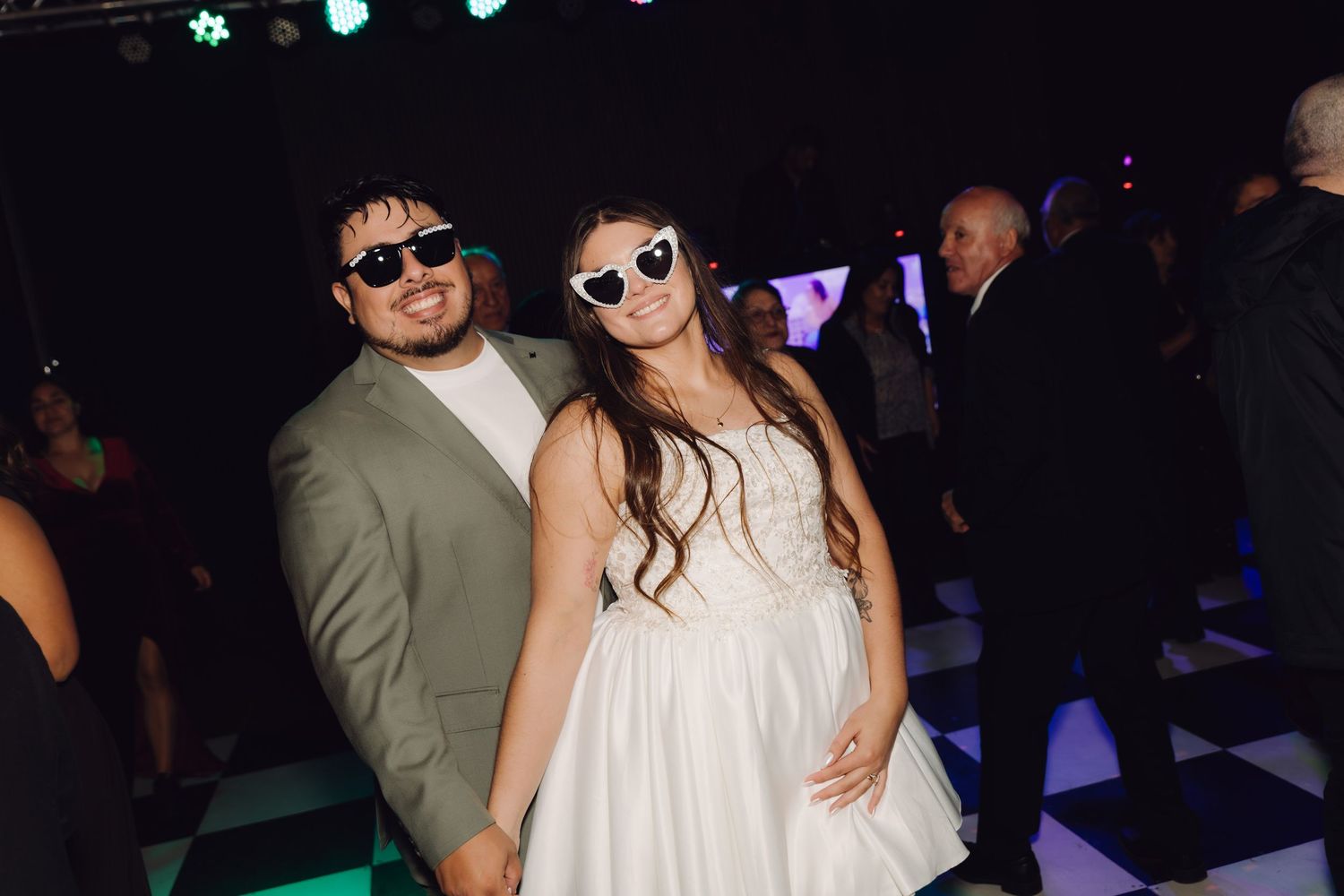 Couple in wedding attire wear white sunglasses on a checkered dance floor.
