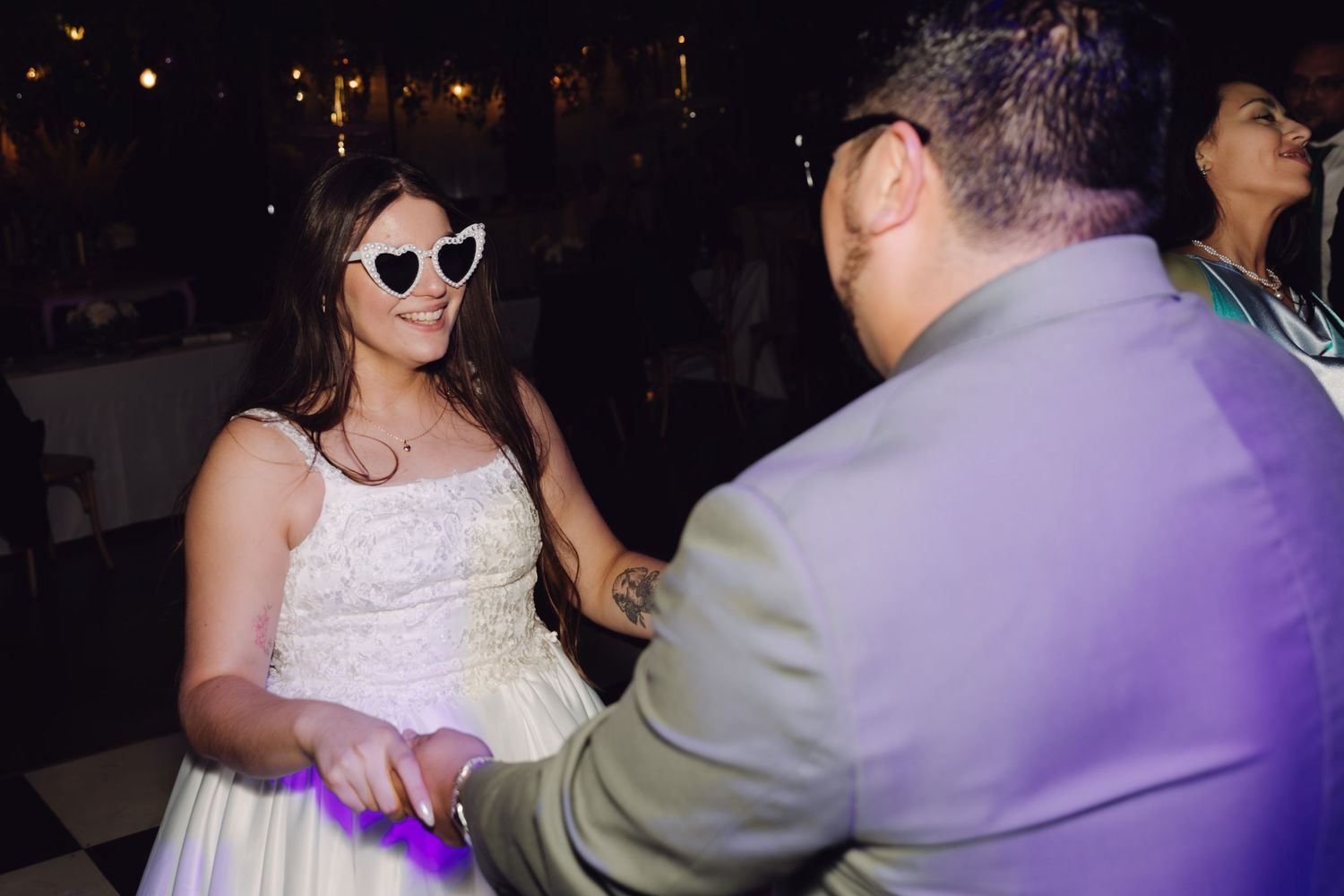 Bride in heart-shaped sunglasses dances joyfully with groom at reception.