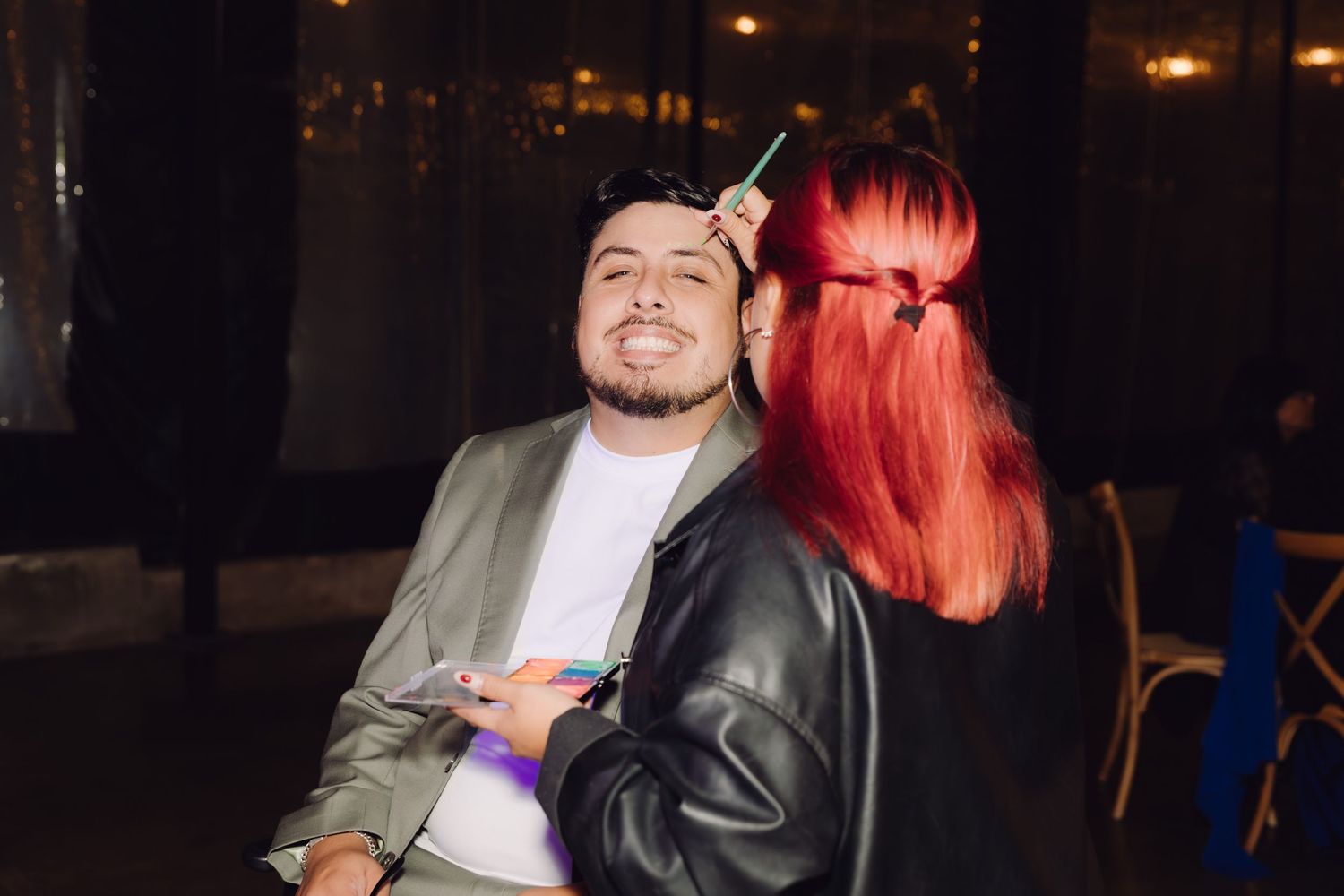 Young man smiling as a red-haired woman playfully holds a unicorn horn to his forehead at a night event.