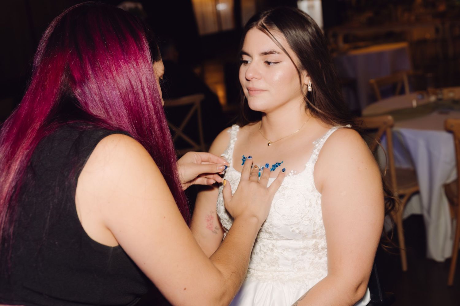 Woman with pink hair adjusting jewelry on a young woman in a white dress at a formal celebration.