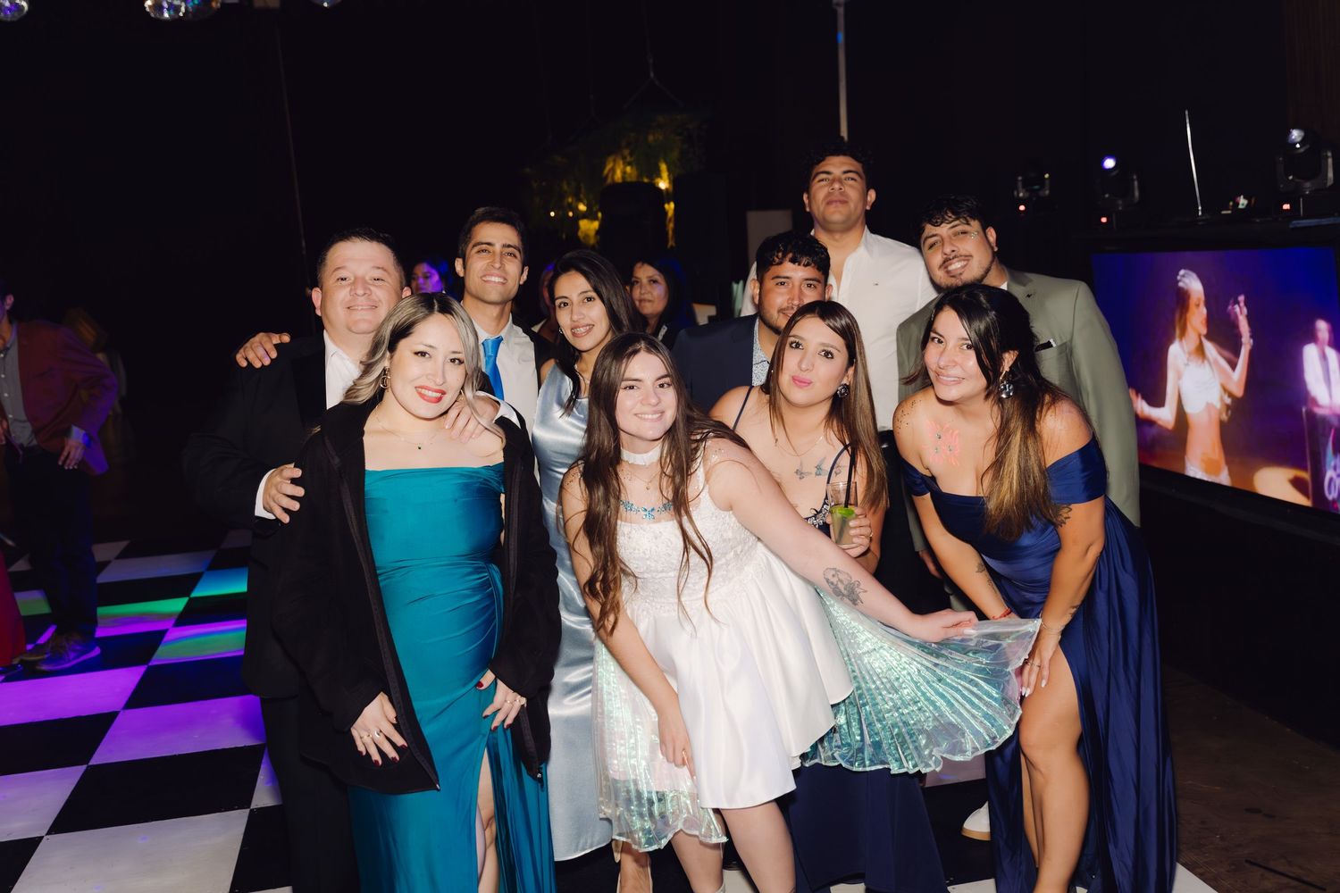 Large group of elegantly dressed young people posing together on a checkered dance floor at night.