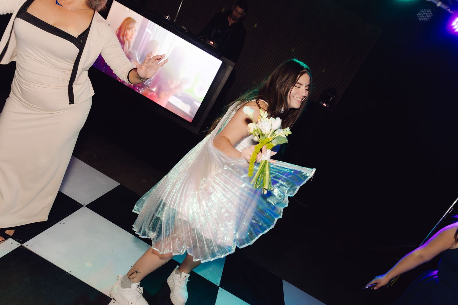 Young girl in a shimmering dress carrying flowers walking across a checkered dance floor at a party.