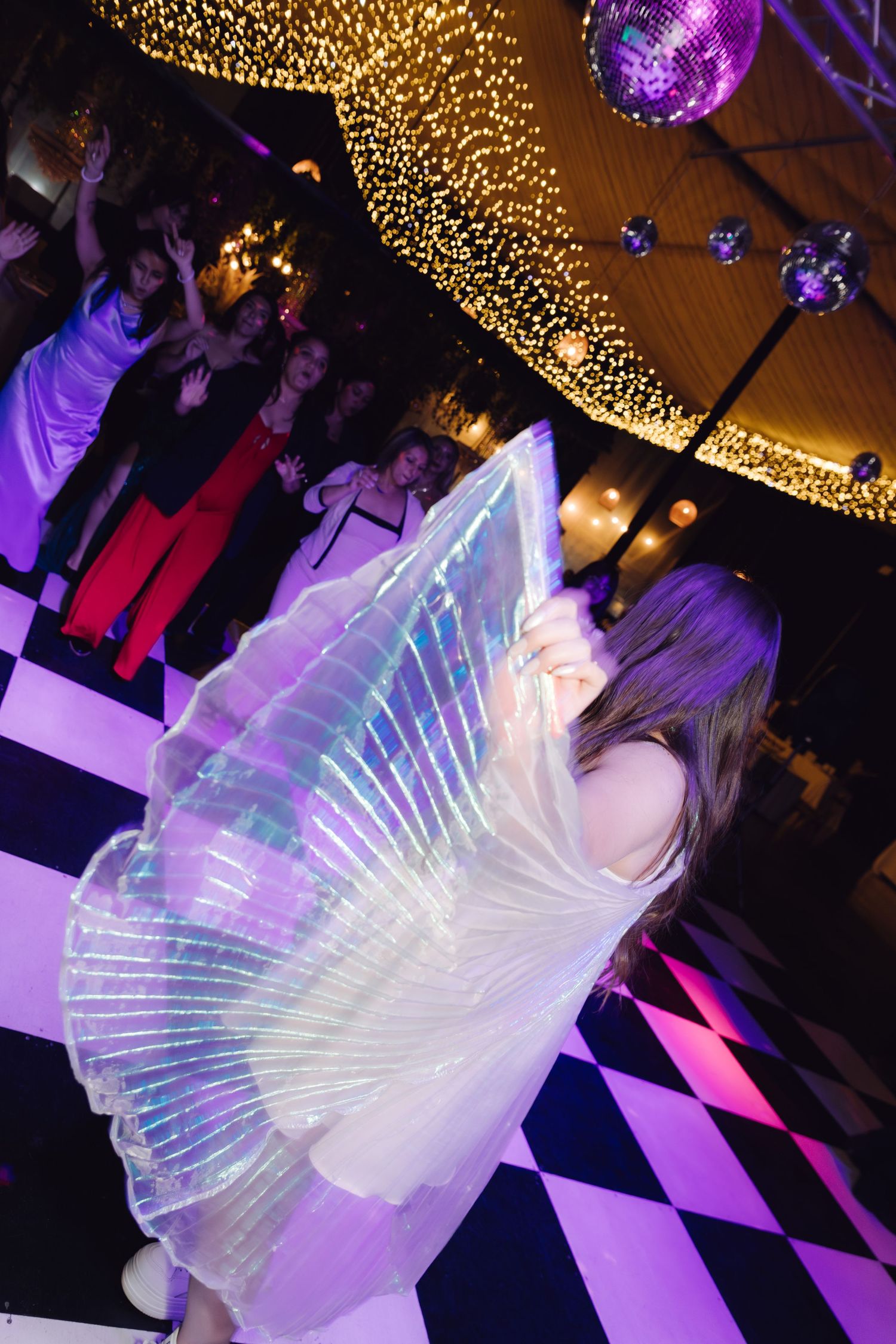 Performer dancing with iridescent fan wings under disco balls on a checkered floor at a gala event.