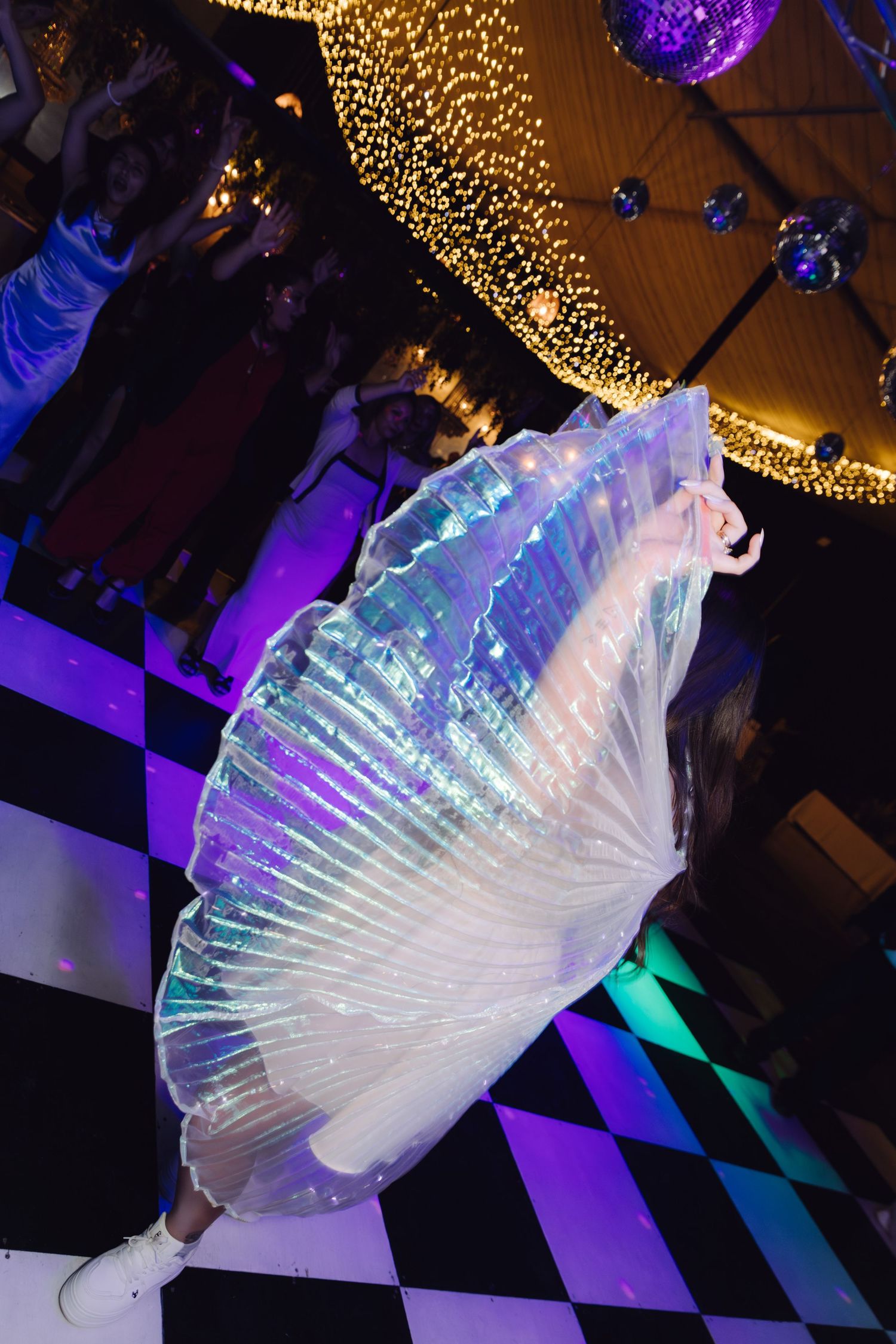 Dancer spinning with large iridescent fan wings on a black and white checkered dance floor at night.
