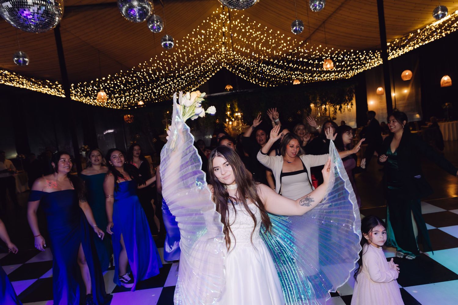 Bride in white gown with iridescent wings dancing at elegant wedding reception with string lights.
