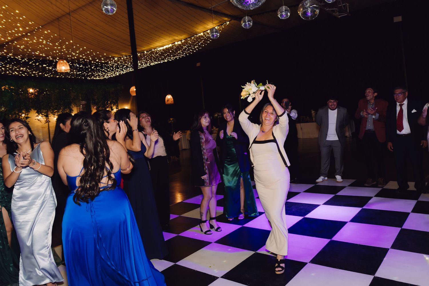 Woman tossing bridal bouquet to guests on checkered dance floor at wedding reception.
