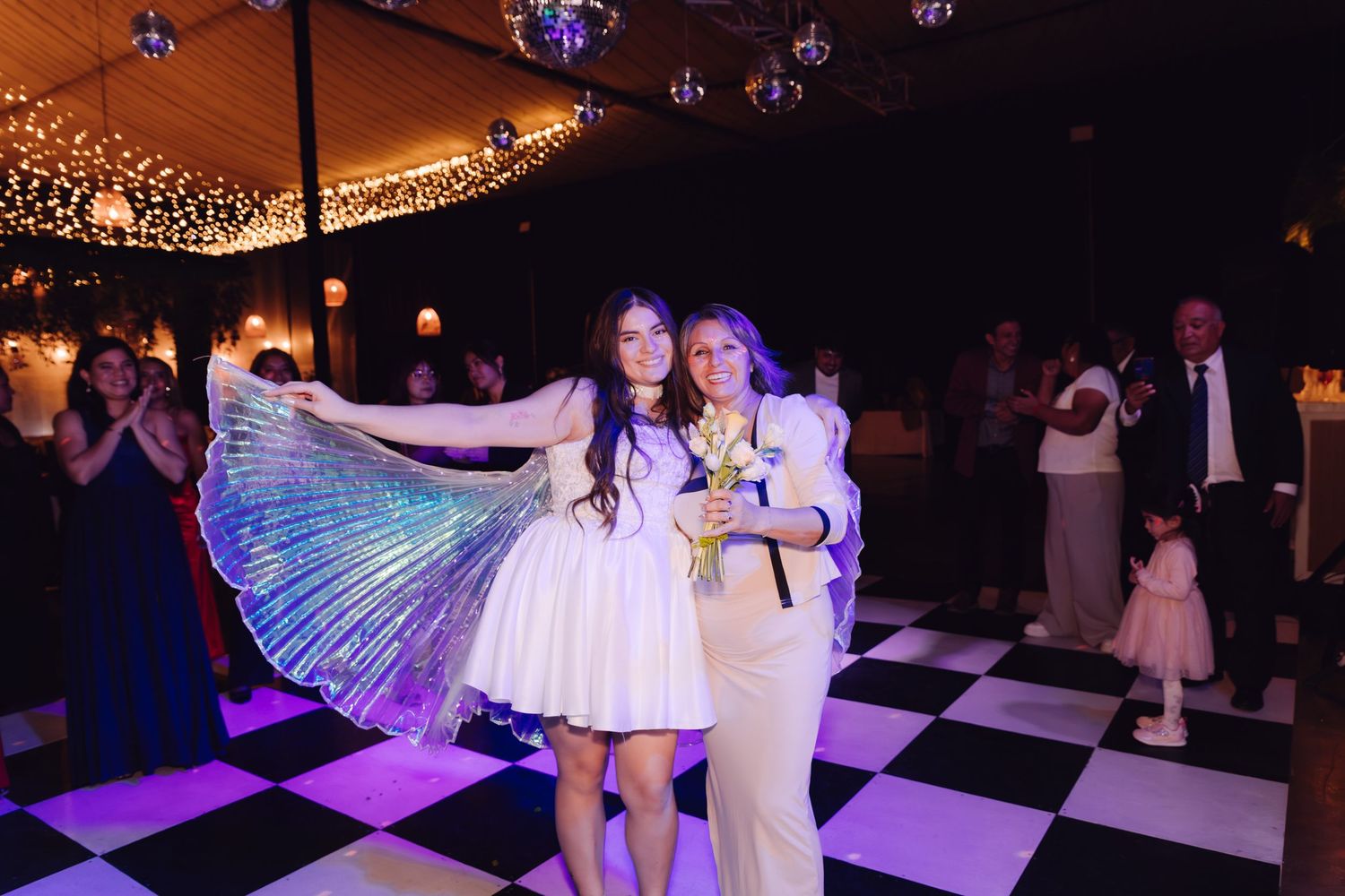 Bride displaying colorful iridescent butterfly wings with woman on black and white dance floor.