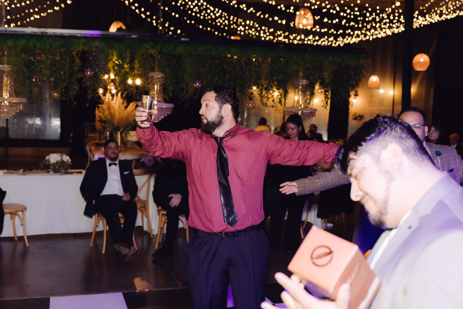 Man in burgundy shirt raising glass in toast at festive indoor wedding celebration.